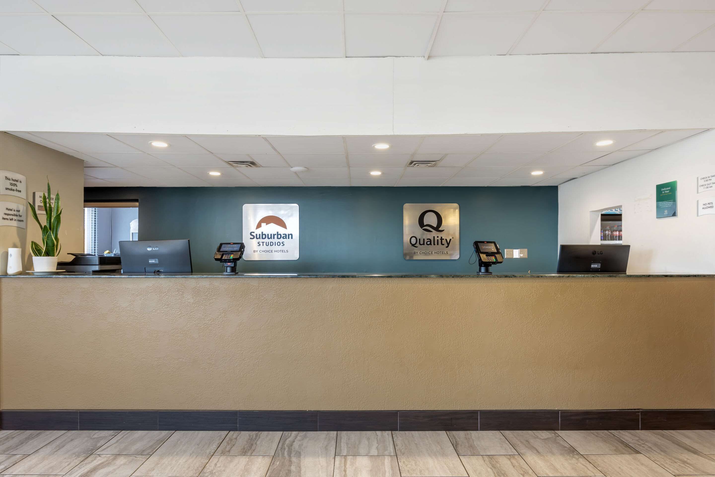 A hotel reception desk with a teal wall, two signs reading "Suburban Studios by Choice Hotels" and "Quality by Choice Hotels", and a computer on the desk.