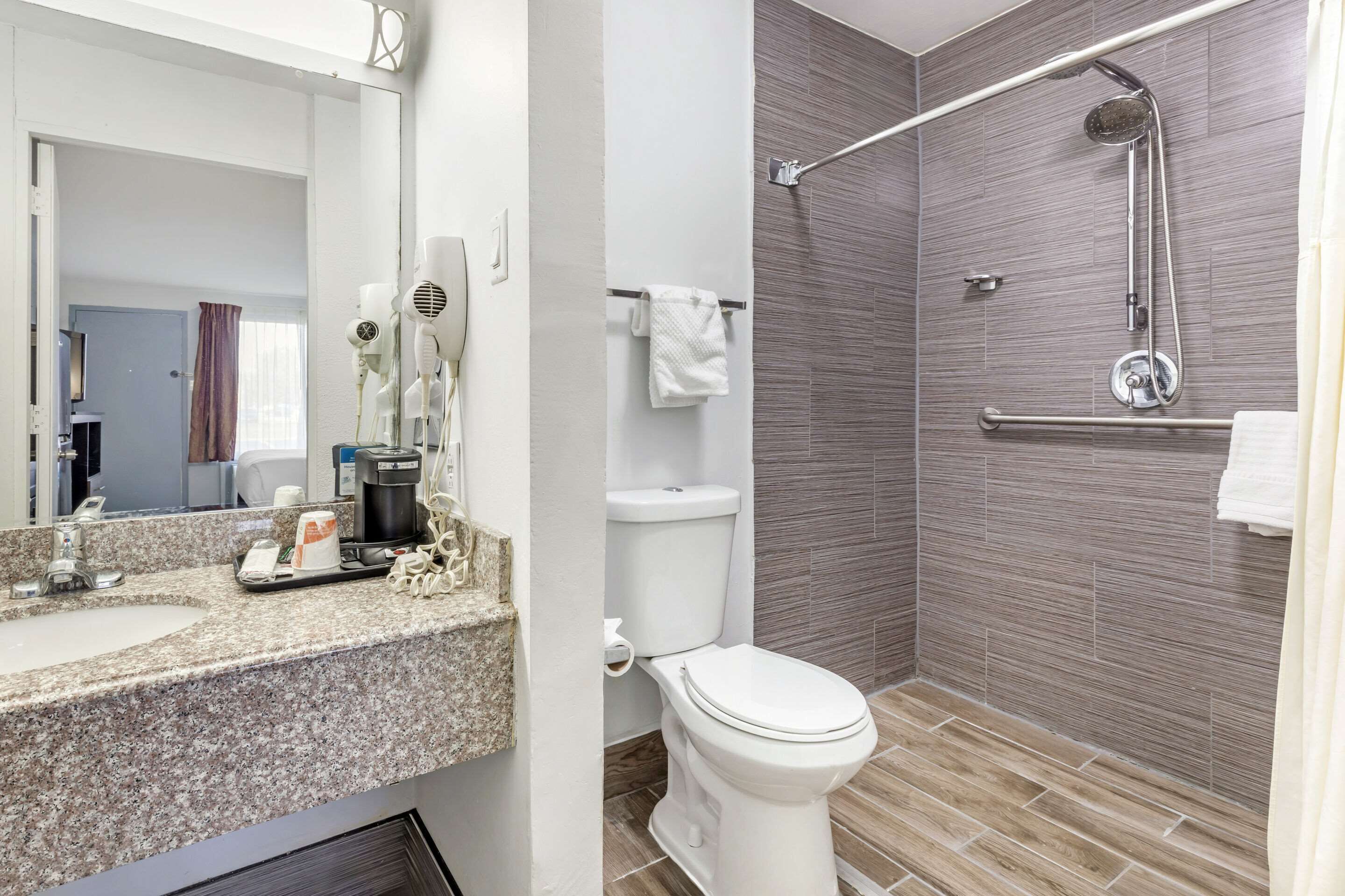 A bathroom with a granite countertop, a white toilet, a shower with a handheld showerhead, and a towel rack.