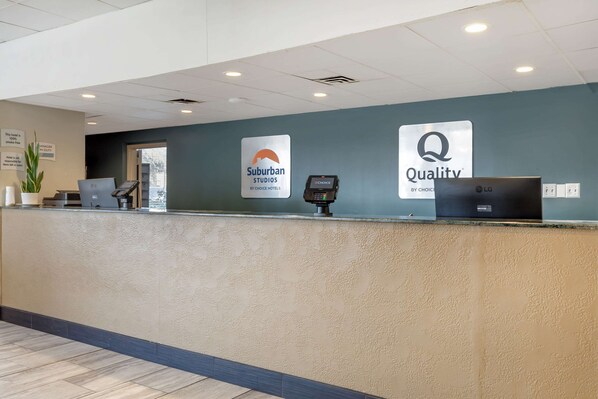 Un mostrador de recepción de hotel con una computadora, un terminal de pago y letreros que dicen 'Suburban Studios by Choice Hotels' y 'Quality by Choice Hotels'.