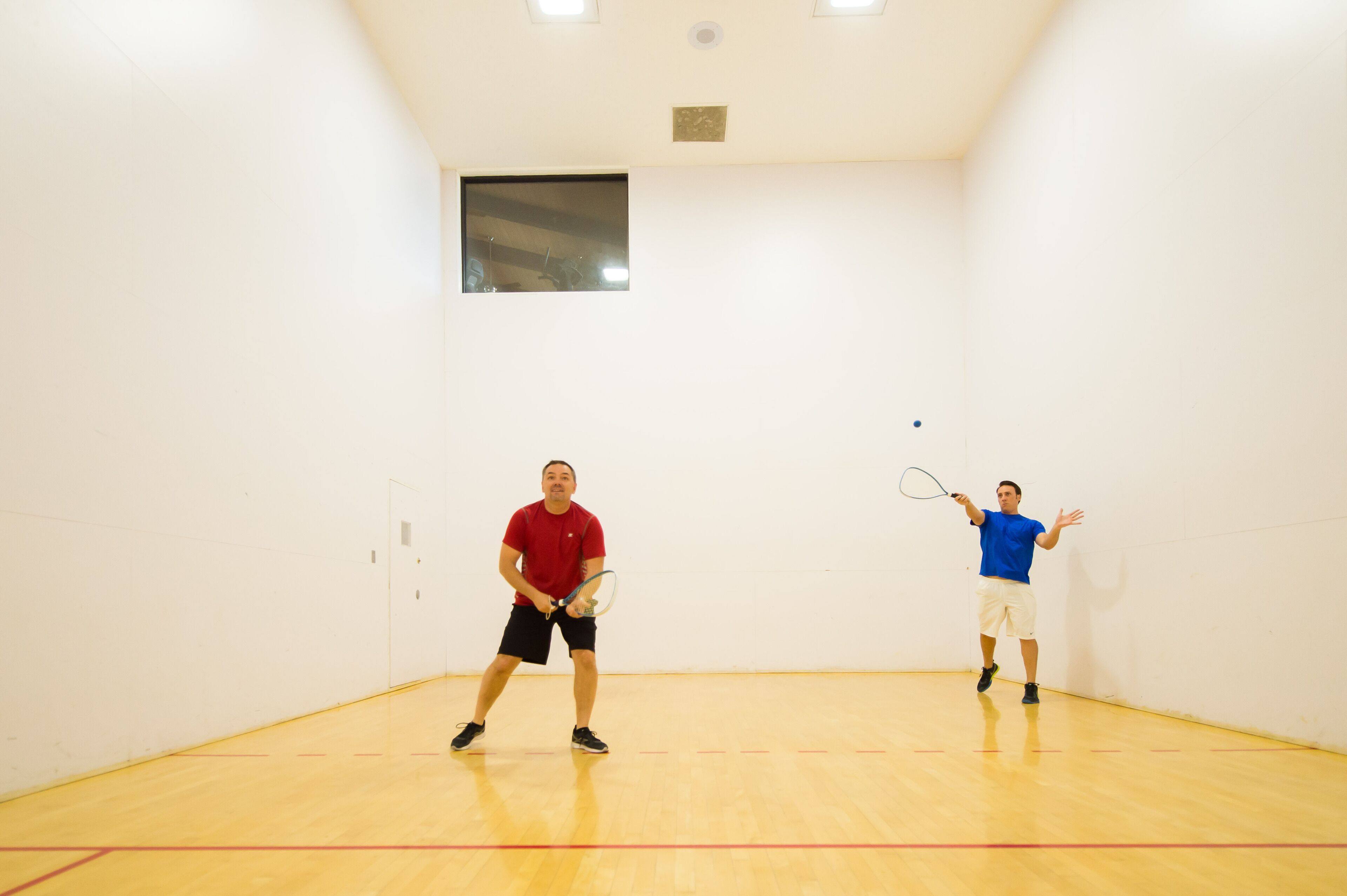Zwei Personen spielen Squash in einem Raum mit Holzboden und weißen Wänden.