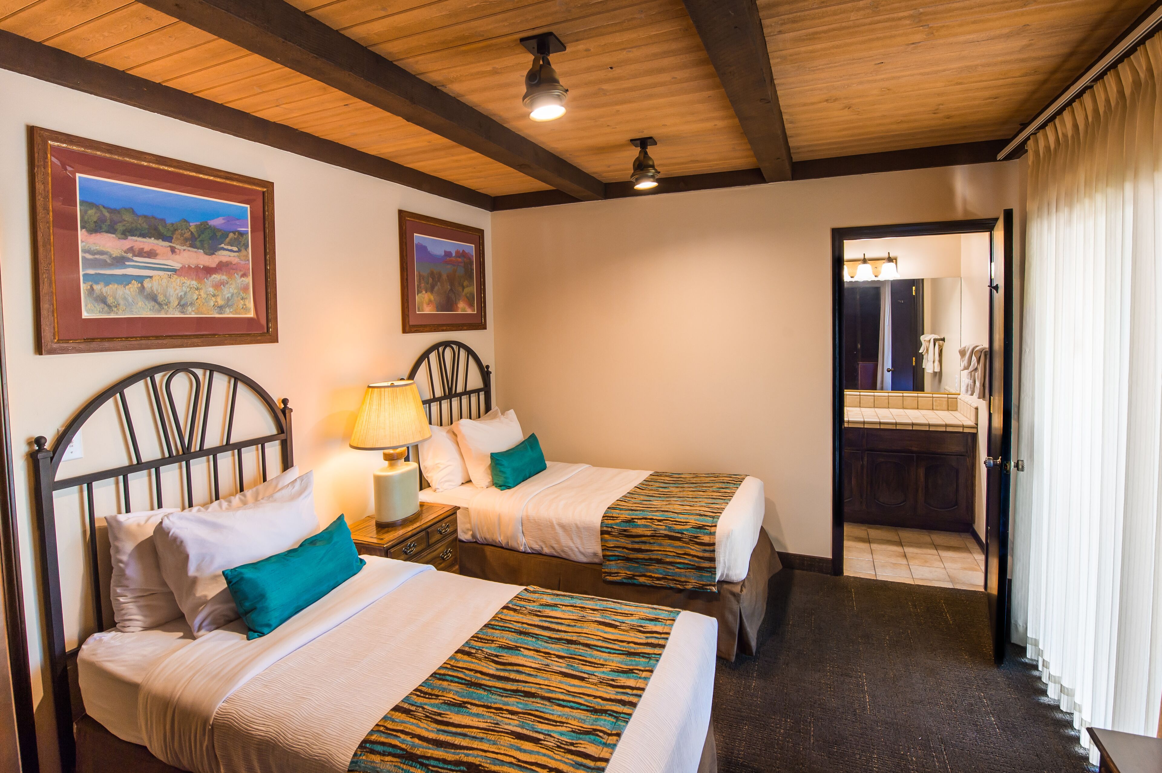 A hotel room with two beds, a wooden ceiling, a nightstand with a lamp, and a bathroom visible through an open door.