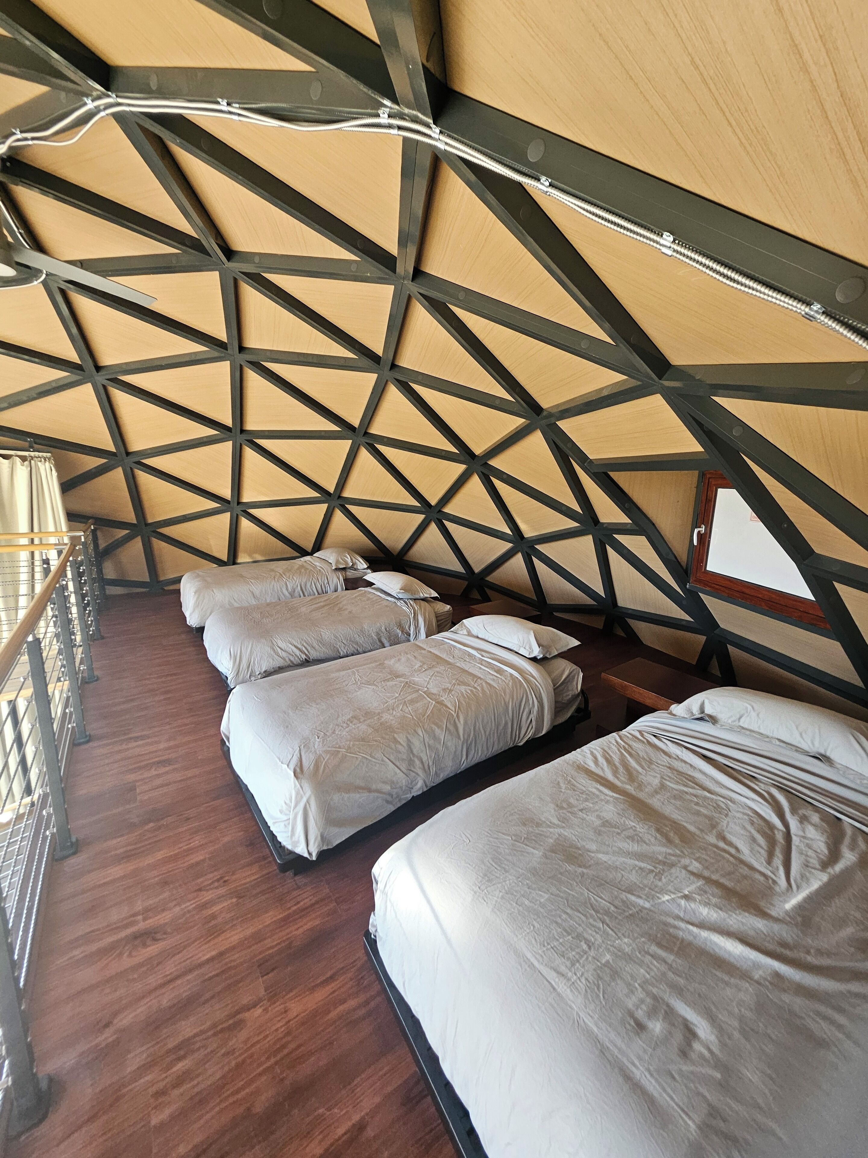 A triangular room with three beds aligned along one wall, wooden flooring, and a metal railing.
