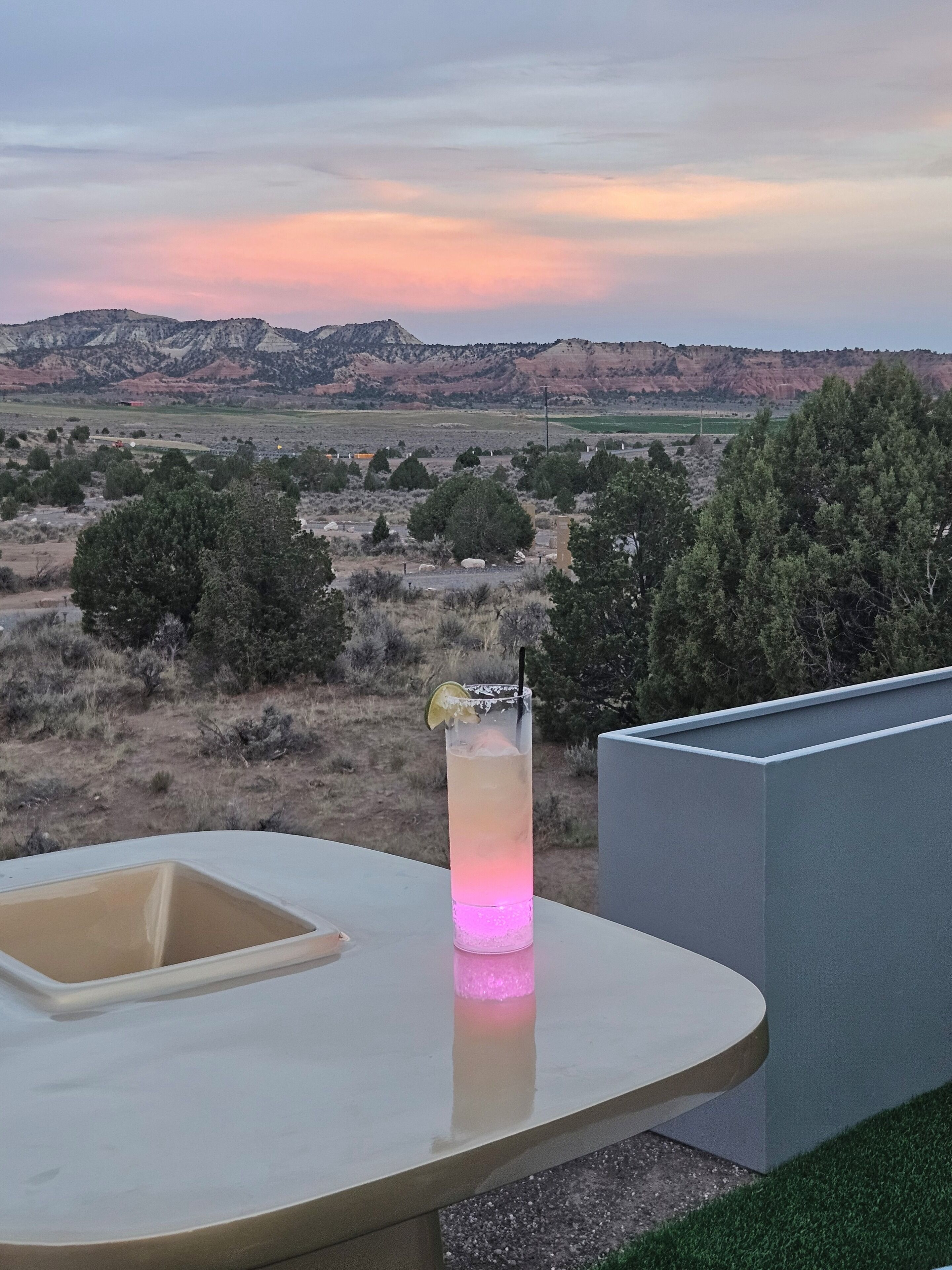 A balcony with a table, a drink with a lime wedge, and a view of a desert landscape with mountains in the distance.