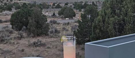 A balcony with a table, a drink with a lime wedge, and a view of a desert landscape with mountains in the distance.