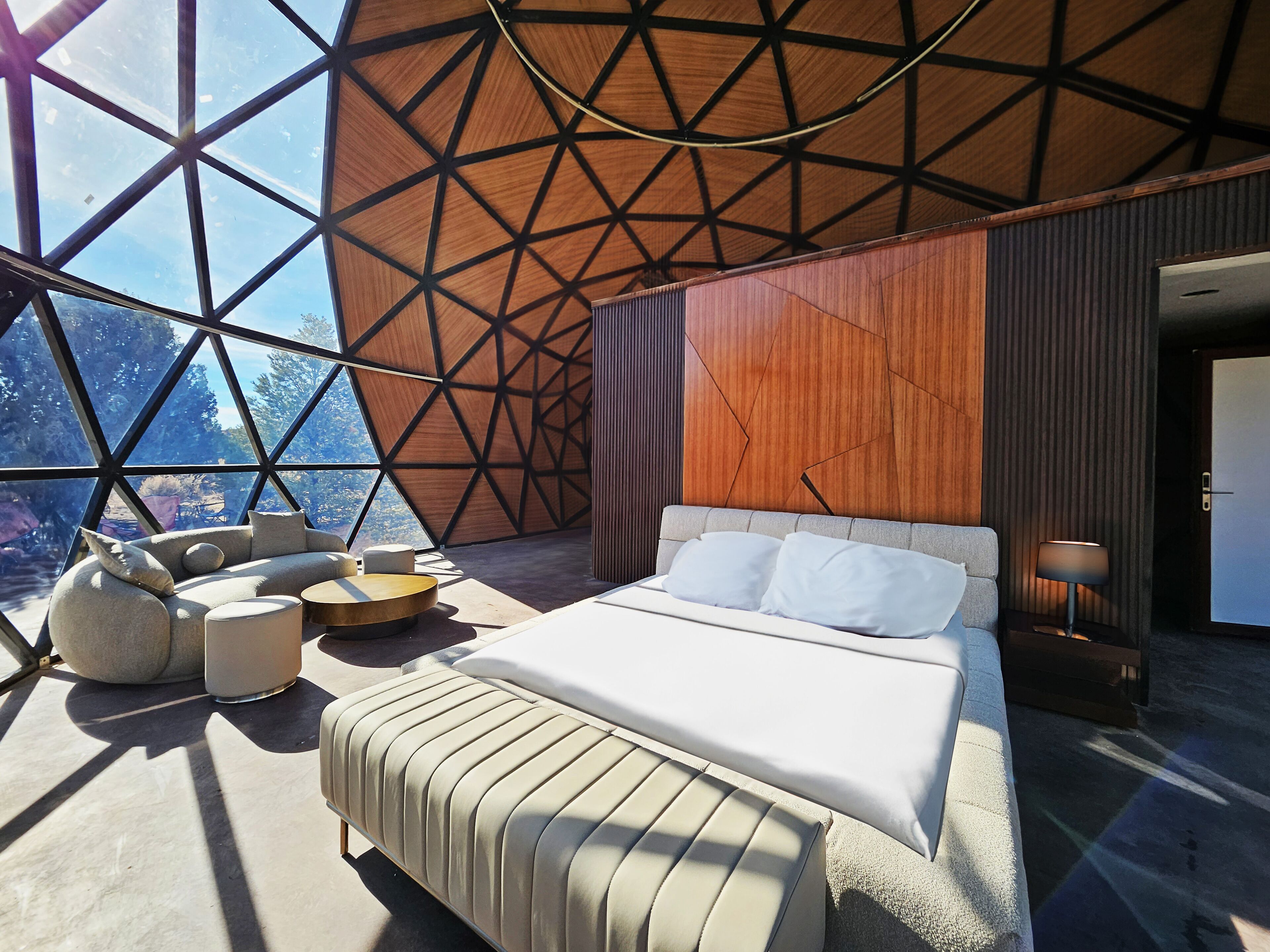 A modern bedroom with a geometric wooden wall, a large bed with white bedding, and a view of the outdoors through large windows.