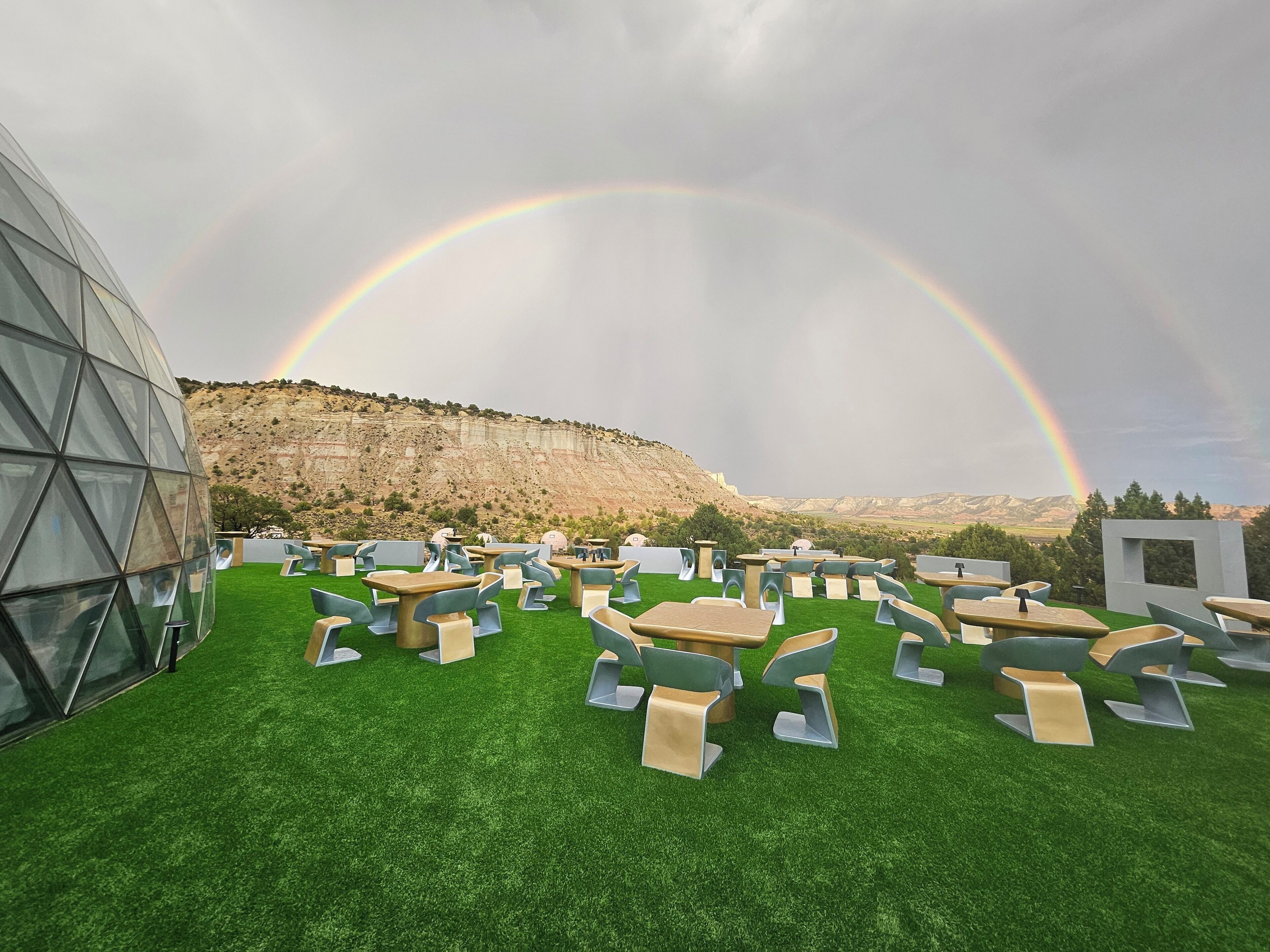An outdoor seating area with tables and chairs on artificial grass, a modern glass building, and a mountainous backdrop with a rainbow.