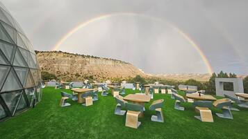An outdoor seating area with tables and chairs on artificial grass, a modern glass building, and a mountainous backdrop with a rainbow.