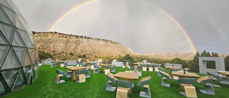 An outdoor seating area with tables and chairs on artificial grass, a modern glass building, and a mountainous backdrop with a rainbow.