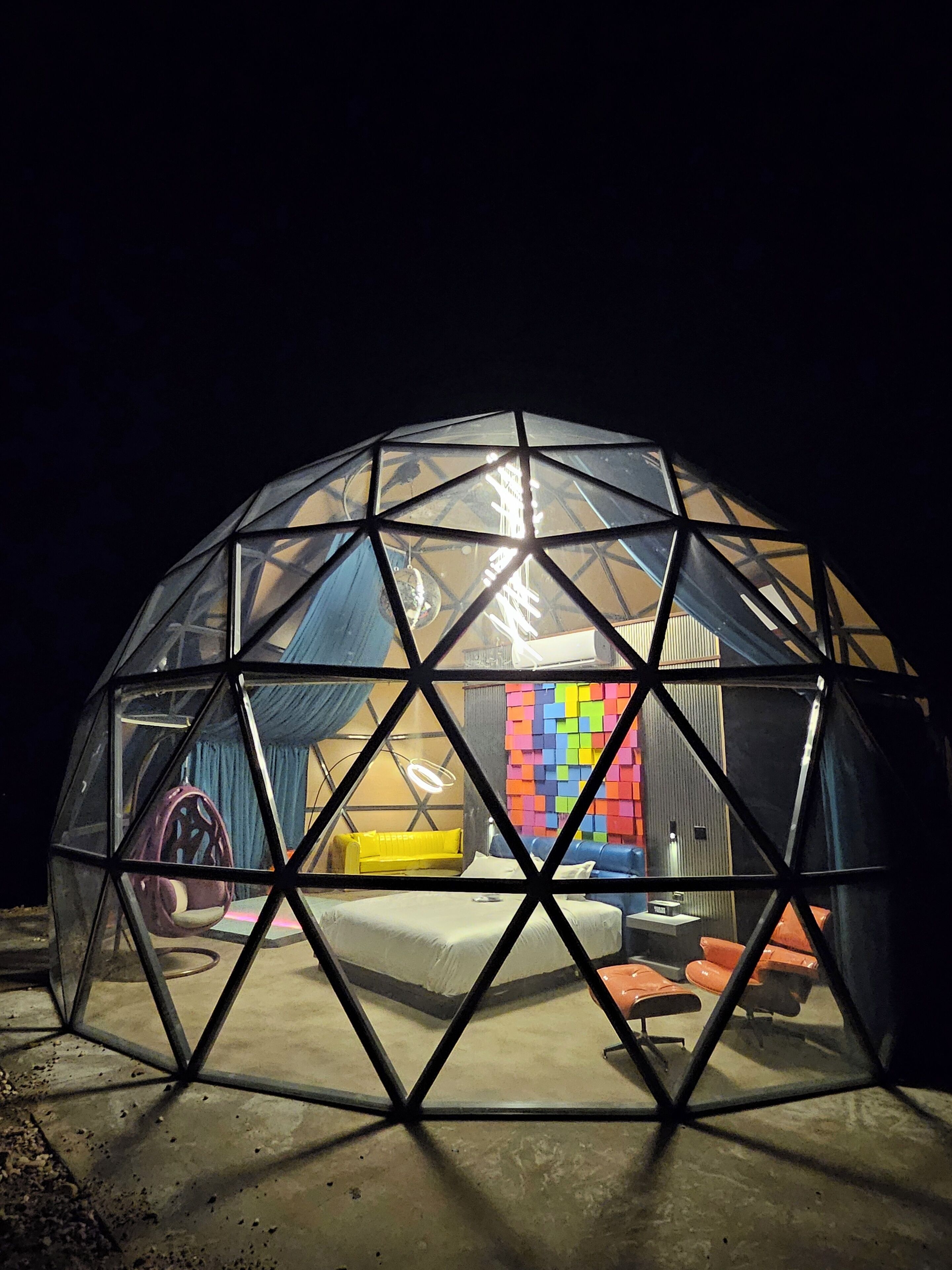 A geodesic dome structure with a transparent roof, revealing a colorful interior with a bed, pillows, and a hanging light fixture.