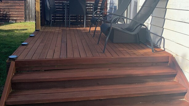 Una terraza de madera con mobiliario exterior, incluyendo sillas y una mesa, al lado de un edificio blanco con una ventana.
