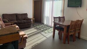 Una sala de estar con un sofá marrón, una mesa de comedor de madera con sillas, una pequeña mesita auxiliar de madera y un televisor montado en la pared.