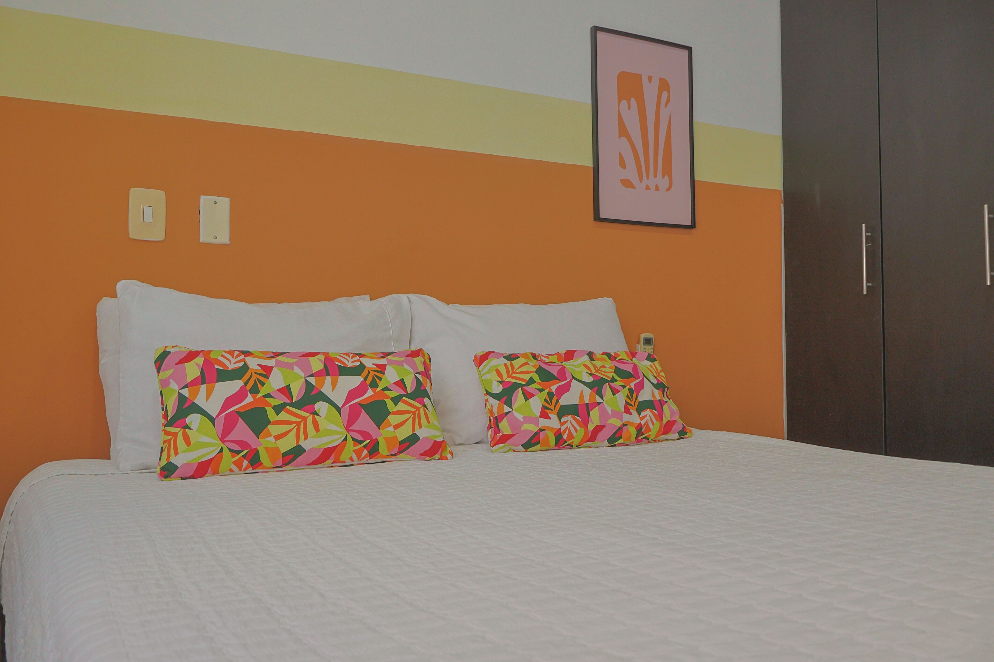 Una cama con ropa blanca, dos almohadas coloridas, un cuadro en la pared y un armario oscuro.