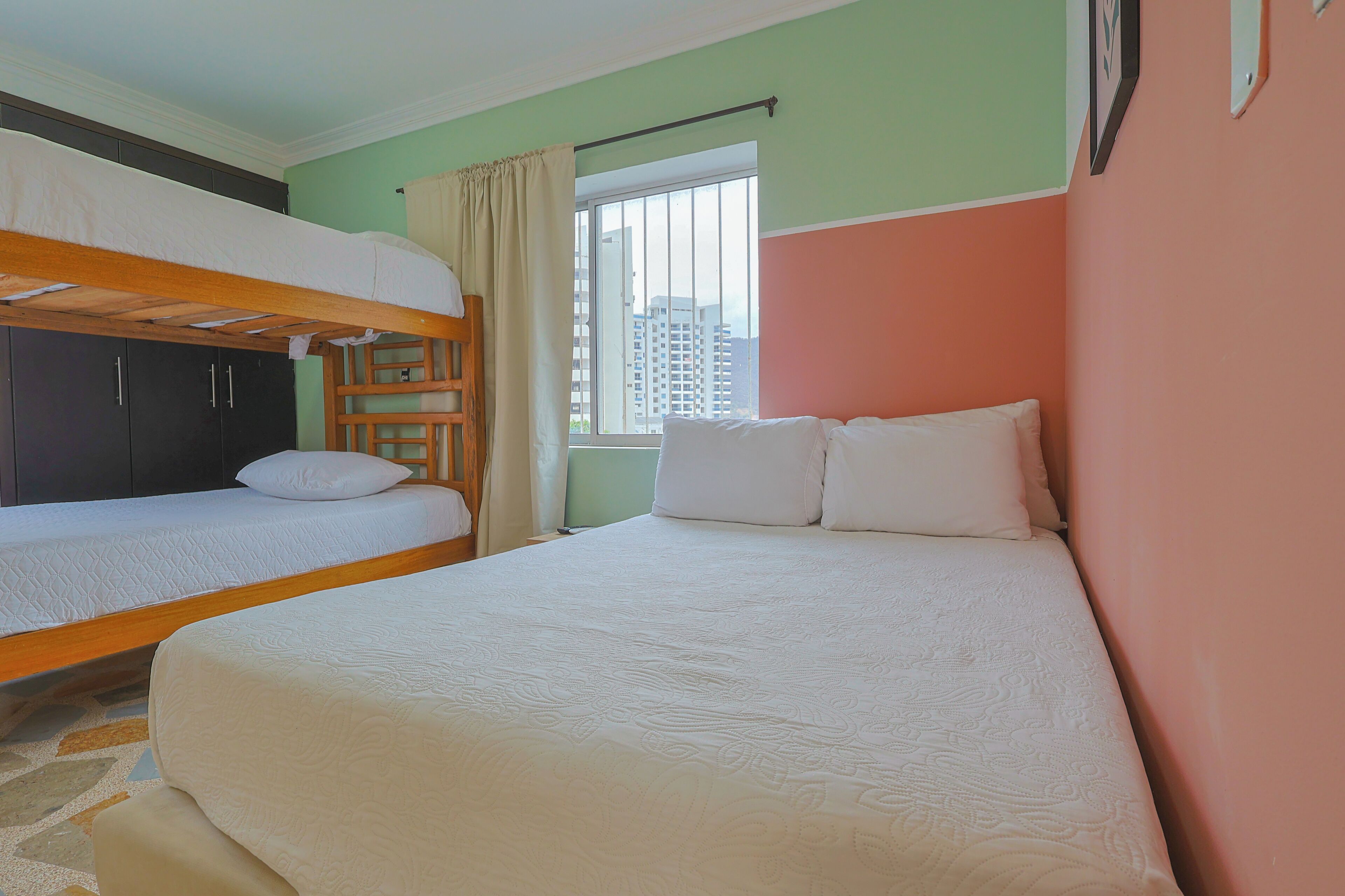 A room with bunk beds, a window with curtains, and a view of buildings outside.
