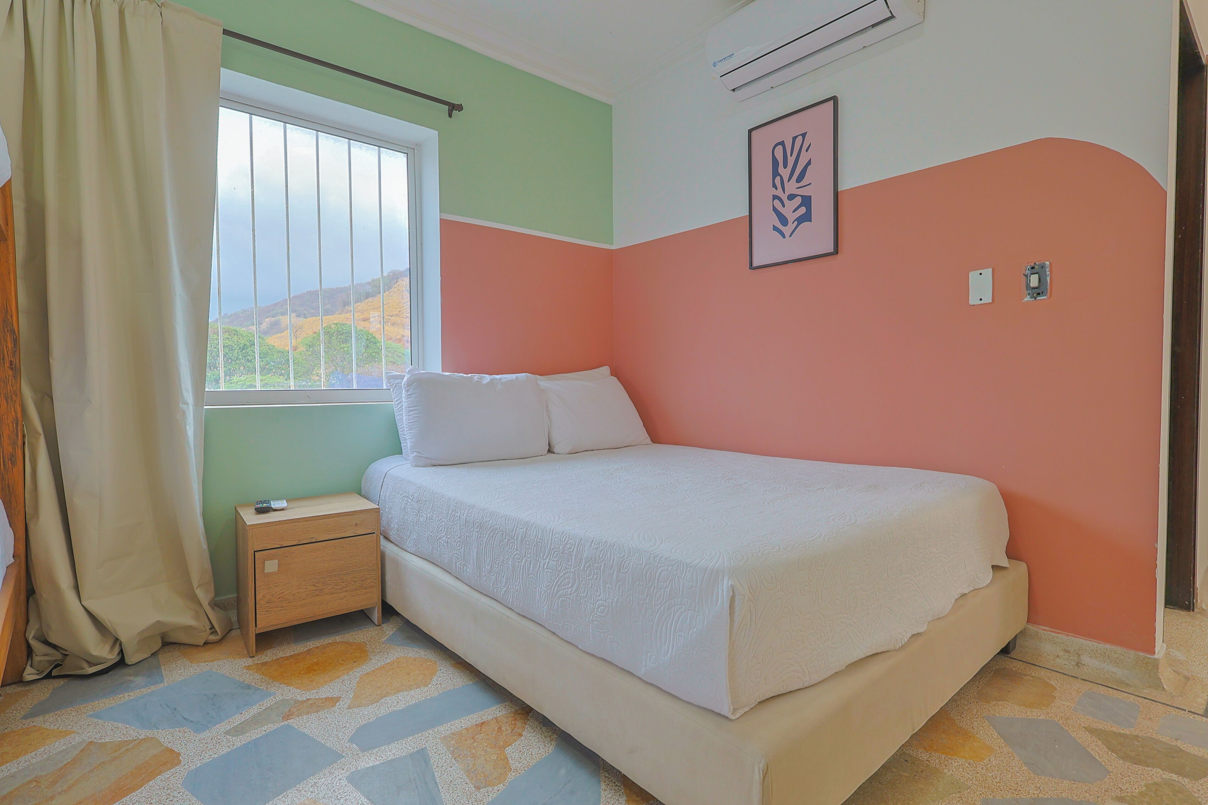 A bedroom with a bed, a nightstand, a window with curtains, and a wall-mounted air conditioner.