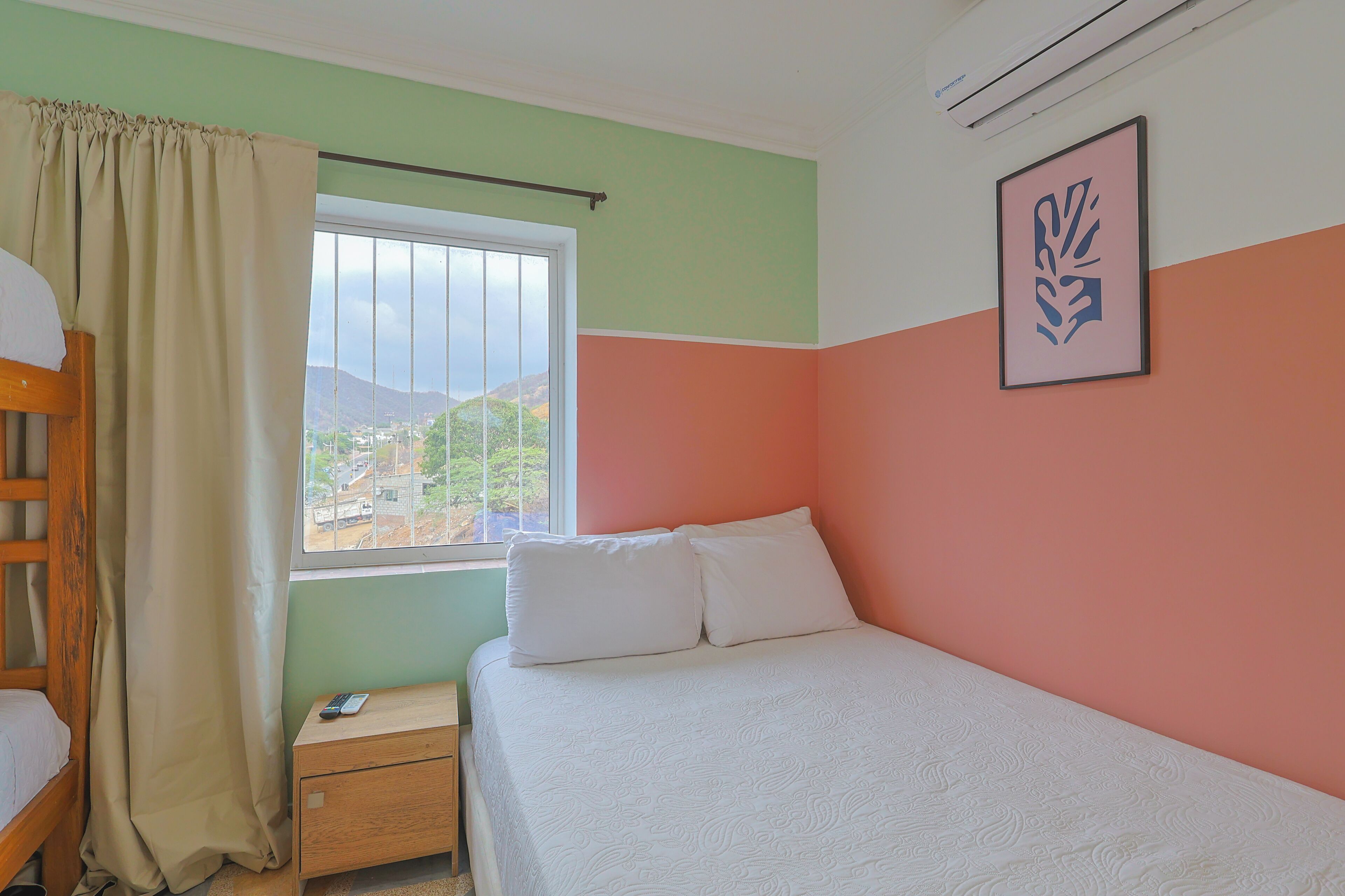 Ein Schlafzimmer mit Bett, Nachttisch, einem Fenster mit Vorhängen und einer wandmontierten Klimaanlage.
