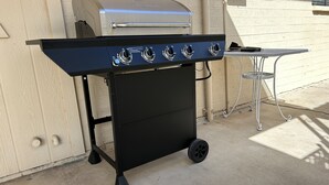 En svart grill med fyra brännare och ett silverfärgat lock, placerad på en betongterrass intill en vit tegelvägg.