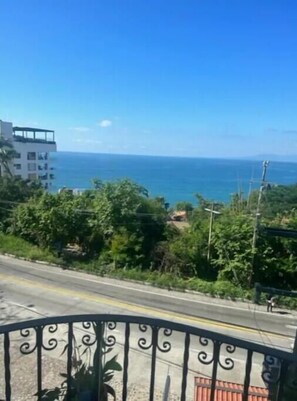 Uma estrada costeira com cerca de metal, vegetação e um edifício com varanda com vista para o mar.