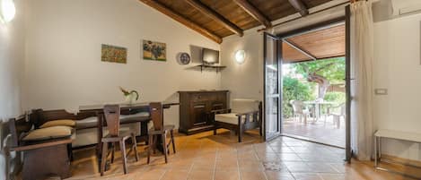A spacious room with a wooden ceiling, tiled floor, a dining area with a table and chairs, a sofa, a cabinet, and a television.