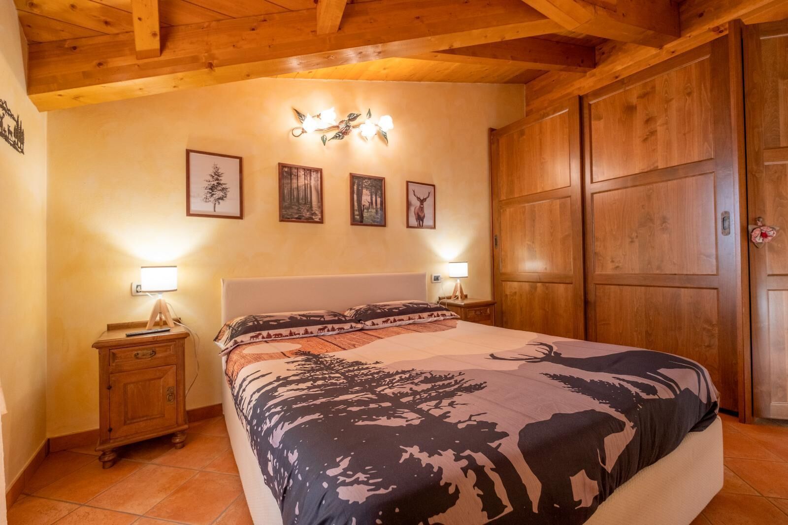 Een slaapkamer met een houten plafond, een bed met een bedsprei met patroon, twee bedlampjes, een nachtkastje en een garderobe.