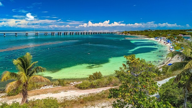 A coastal view with a pier extending into turquoise waters, surrounded by lush greenery and a sandy beach with umbrellas. - Luxury Boating Paradise+Pool+Sailing Kayaks (Marathon)