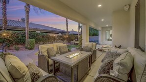 A modern outdoor living area with wicker furniture, a fire feature, and a view of a house in the background. - Serenity: Pool,Spa, Gated Community and Fun! (La Quinta)