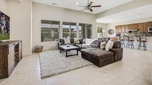 A spacious living room with a large sofa, a coffee table, and a TV. There is a kitchen area with bar stools and a ceiling fan. - Serenity: Pool,Spa, Gated Community and Fun! (La Quinta)