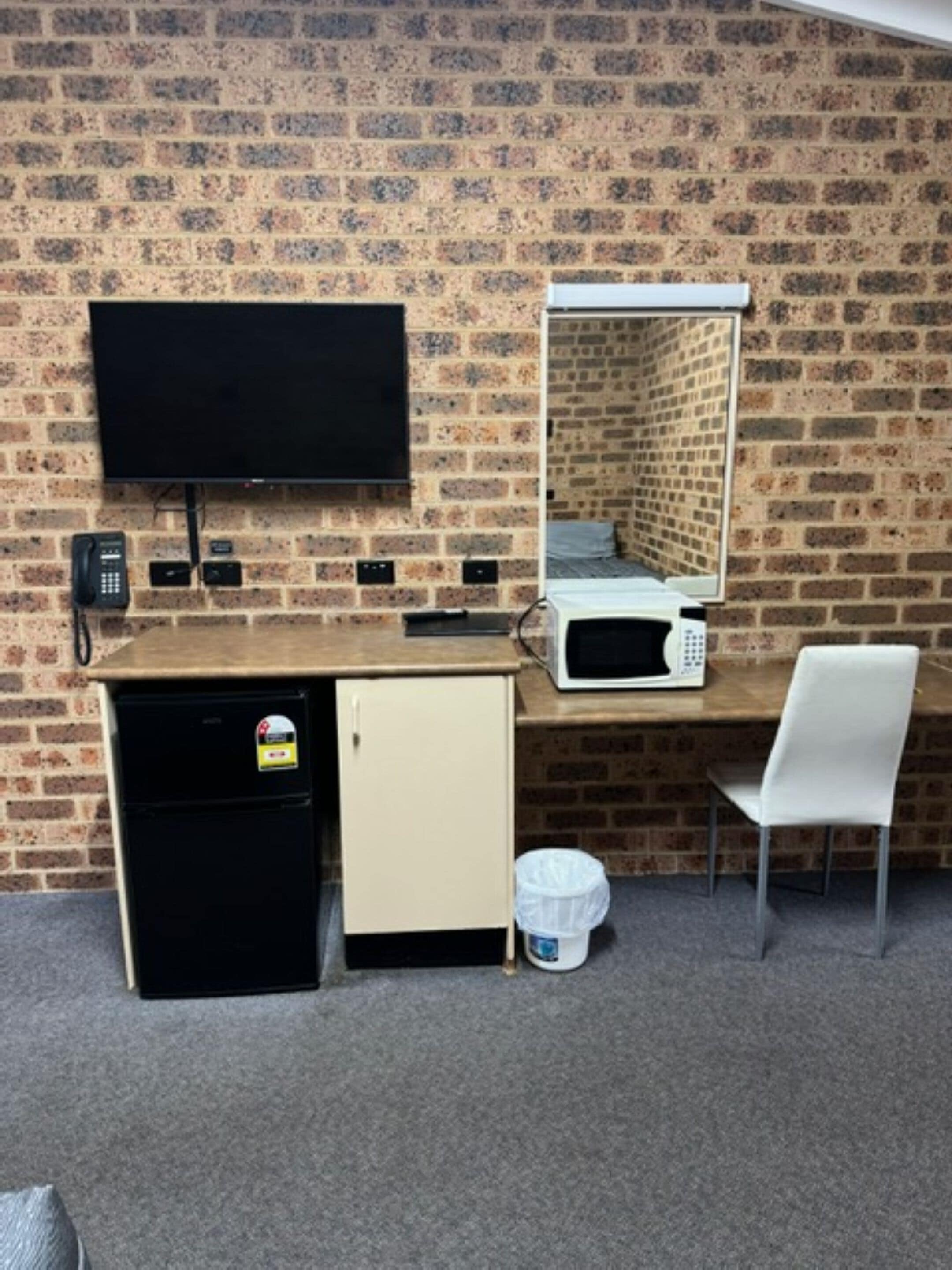Une pièce avec un mur en briques, un téléviseur à écran plat fixé au-dessus d’un comptoir en bois, un four à micro-ondes, un petit réfrigérateur, une chaise blanche et une poubelle.