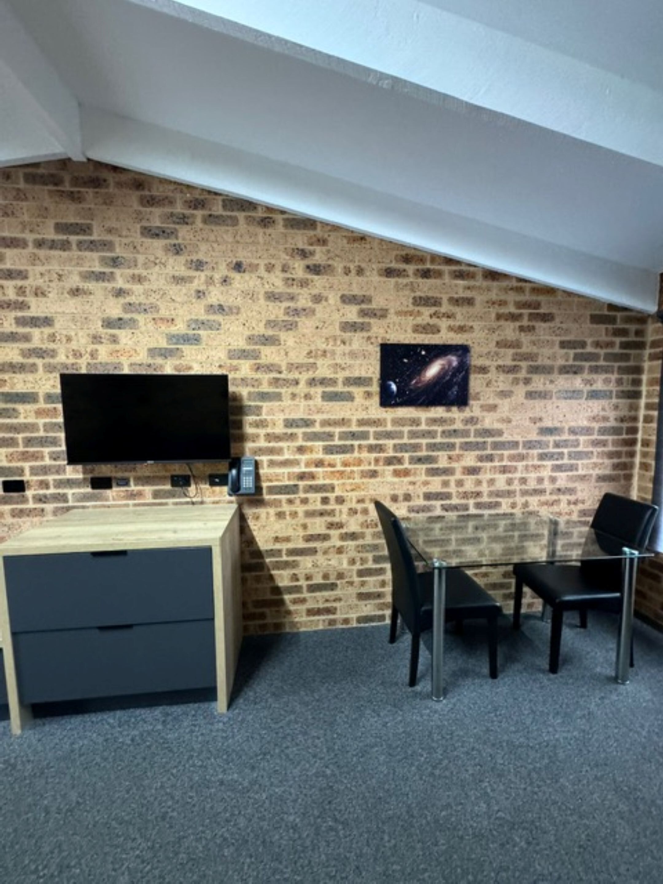 A room with a brick wall, a mounted TV, a desk with drawers, and two chairs.