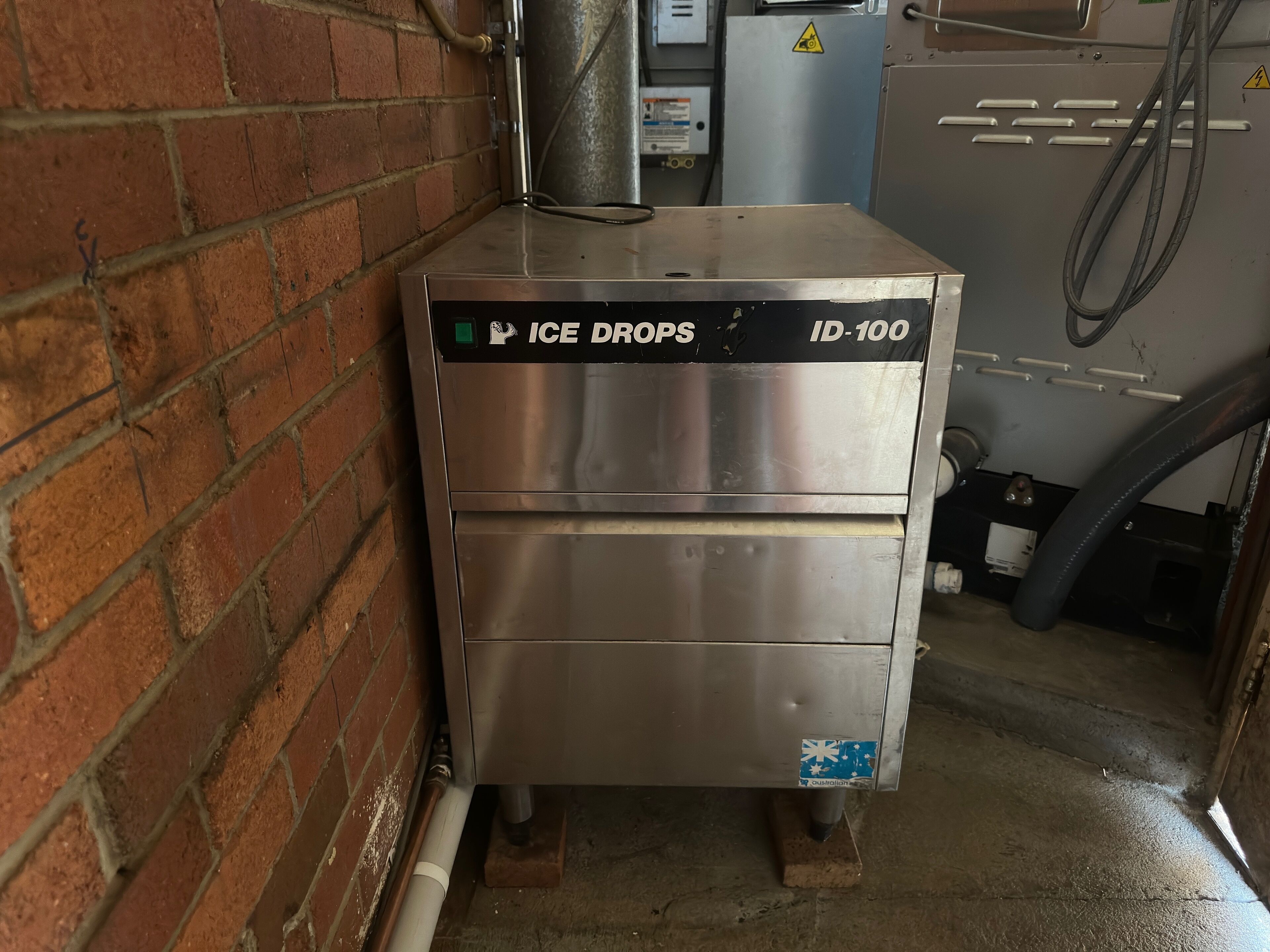 A stainless steel ice machine labeled "ICE DROPS ID-100" in a commercial kitchen with brick walls and a concrete floor.