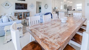 A spacious living room with a large wooden dining table, white chairs, a blue sofa, and a TV mounted on the wall.