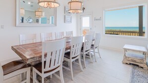 A dining area with a wooden table and white chairs, a chandelier, and a large window with a view of the beach.