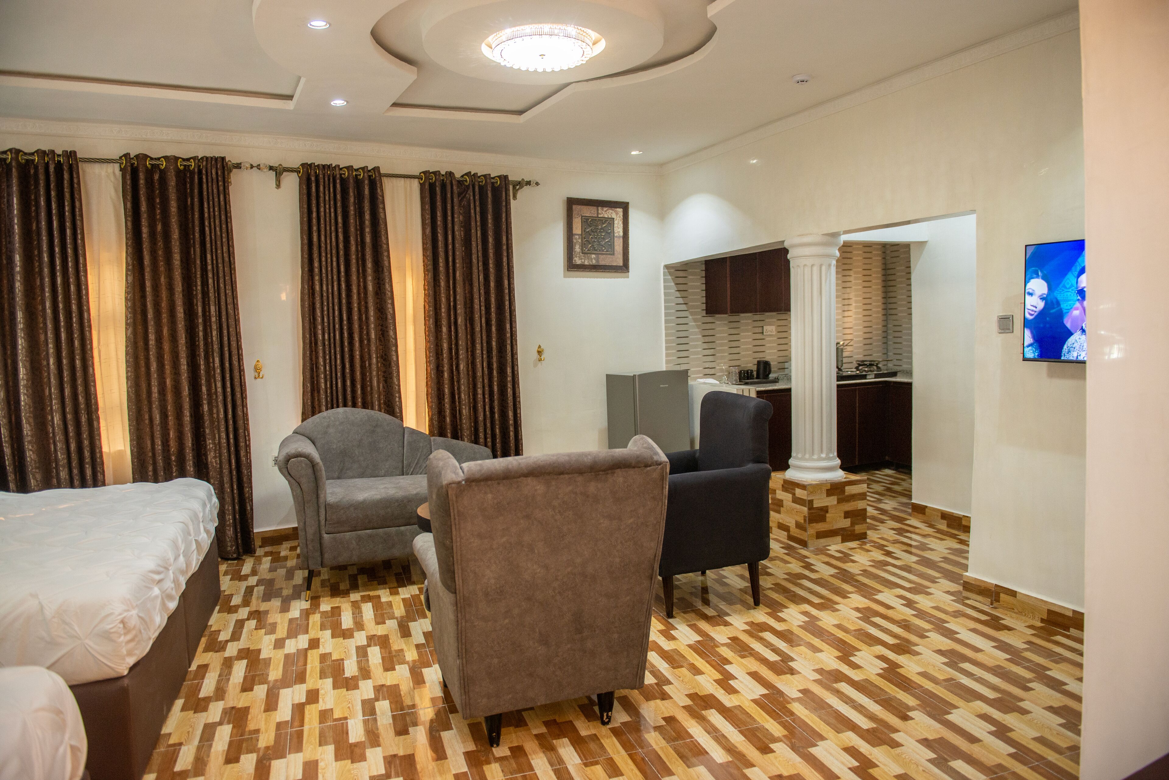 A hotel room with a checkered floor, two armchairs, a bed, a television, and a view of a kitchen area.