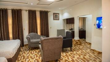 A hotel room with a checkered floor, two armchairs, a bed, a television, and a view of a kitchen area.