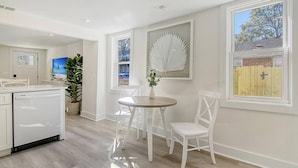 A modern kitchen with white cabinetry, a dishwasher, a dining area with a round table and white chairs, and a large window with a view of a wooden fence. - Renovated - Old Village - Close to Beach/Downtown (Mount Pleasant)