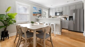 Una cocina moderna con una isla central, muebles blancos, electrodomésticos de acero inoxidable y un comedor con mesa y sillas de madera.