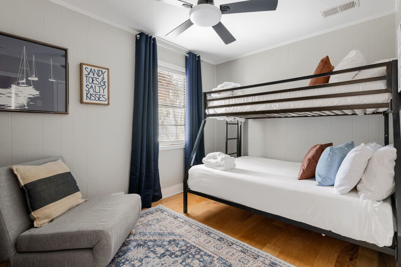 Een slaapkamer met een stapelbed, een grijze bank, een kamerkleed met een patroon en een plafondventilator.