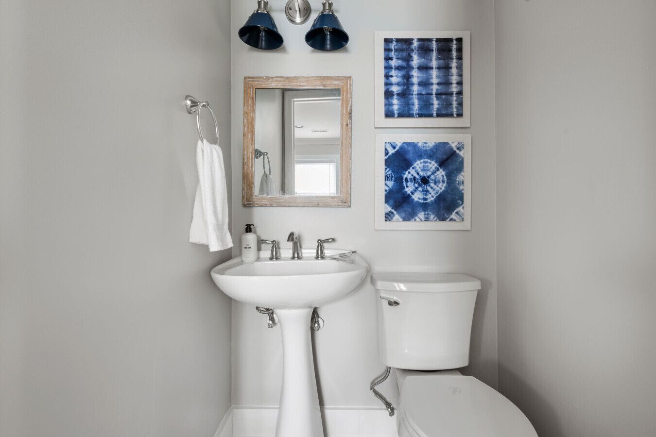 Un baño con un lavamanos blanco, un inodoro y un espejo. Hay dos luces empotradas sobre el lavamanos y dos obras de arte azules enmarcadas en la pared.