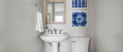 Un baño con un lavamanos blanco, un inodoro y un espejo. Hay dos luces empotradas sobre el lavamanos y dos obras de arte azules enmarcadas en la pared.