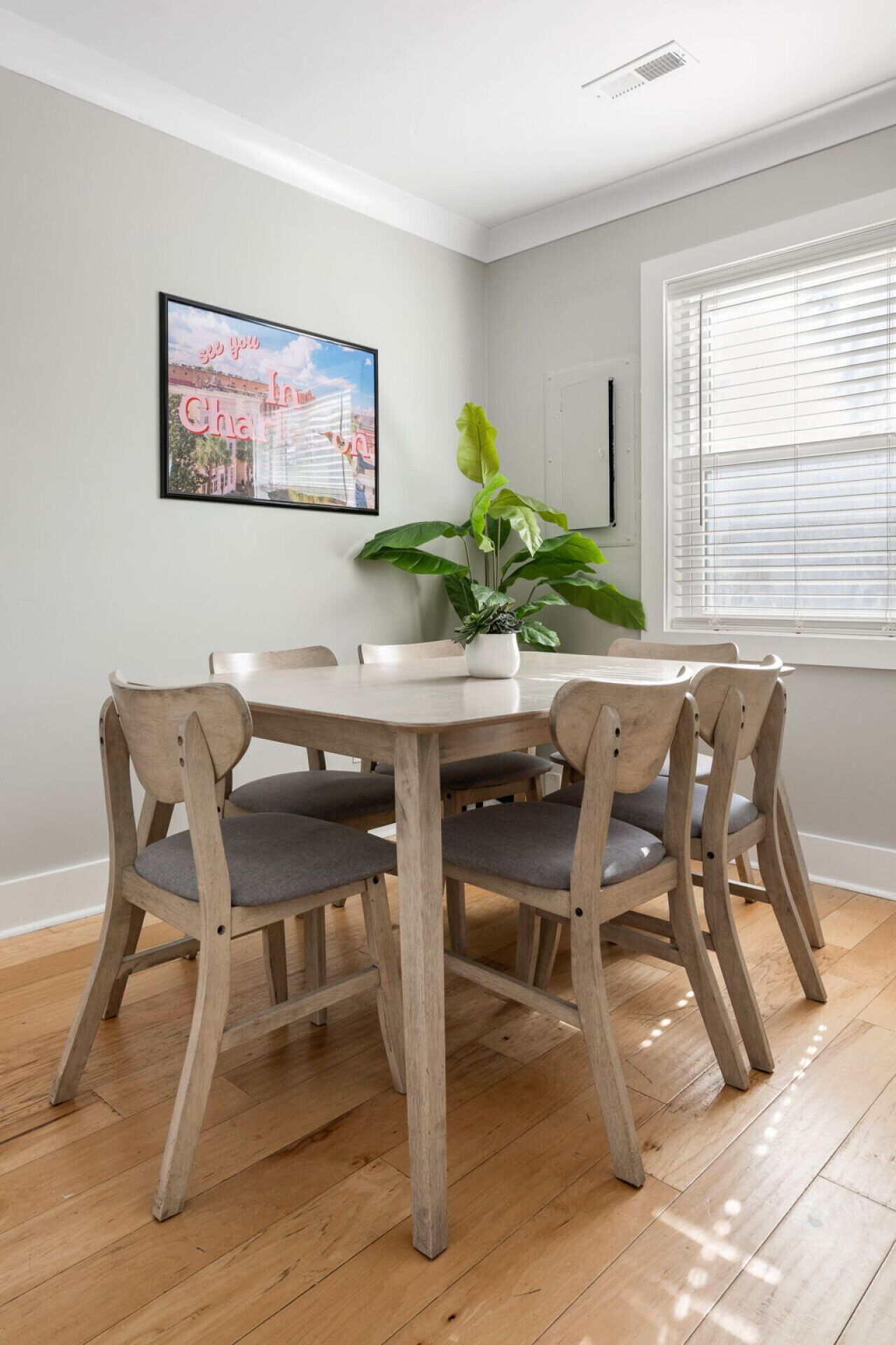 A dining area with a round table and four wooden chairs, a potted plant, and a framed picture on the wall.