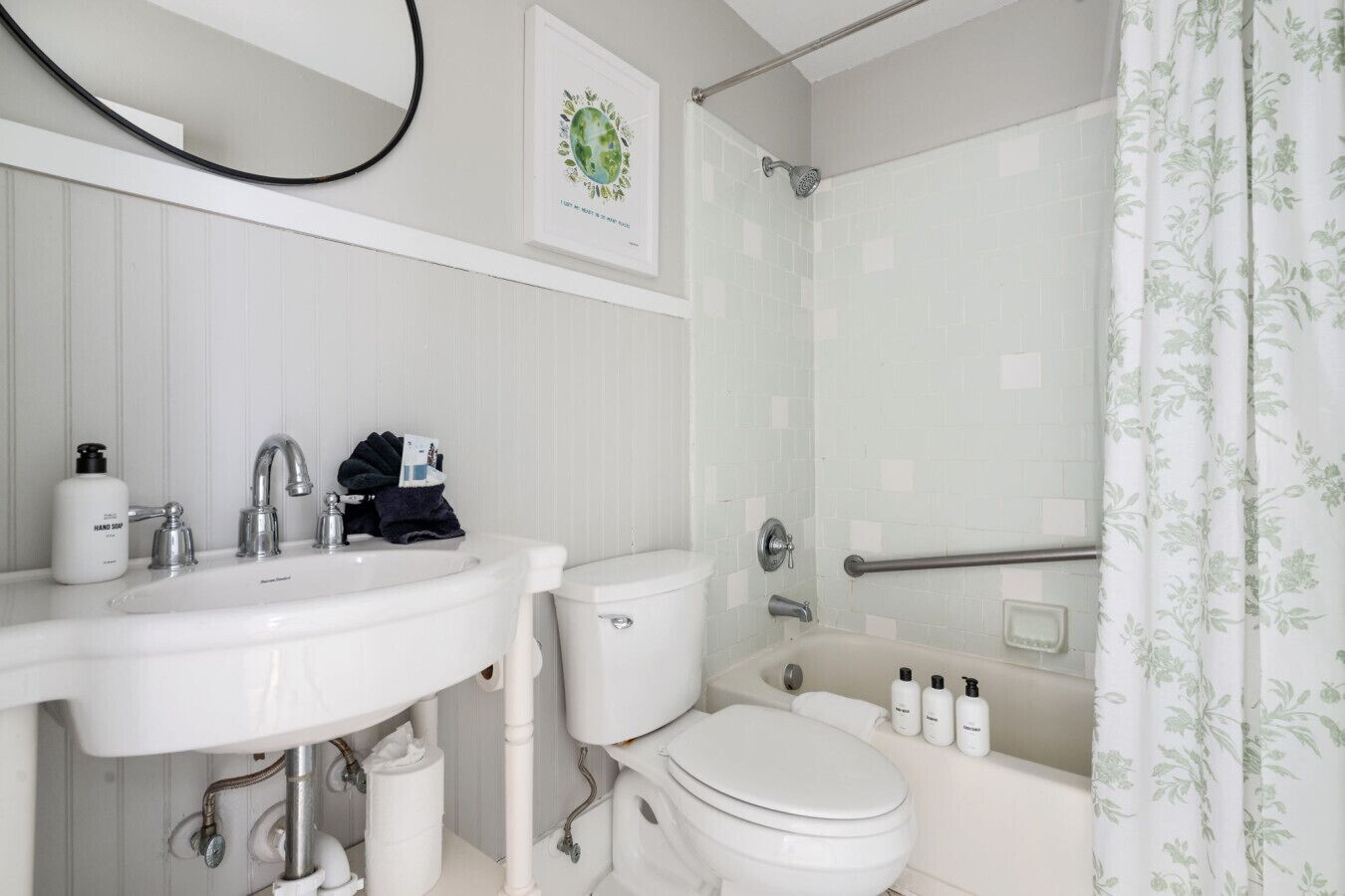 A bathroom with a white sink, toilet, and bathtub. There is a shower curtain with a floral pattern, and a framed picture on the wall.