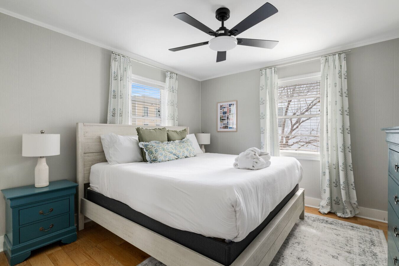 A bedroom with a bed, a nightstand, a dresser, a ceiling fan, and a window with curtains.