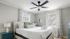 Un dormitorio con cama, mesita de noche, cómoda, ventilador de techo y ventana con cortinas.