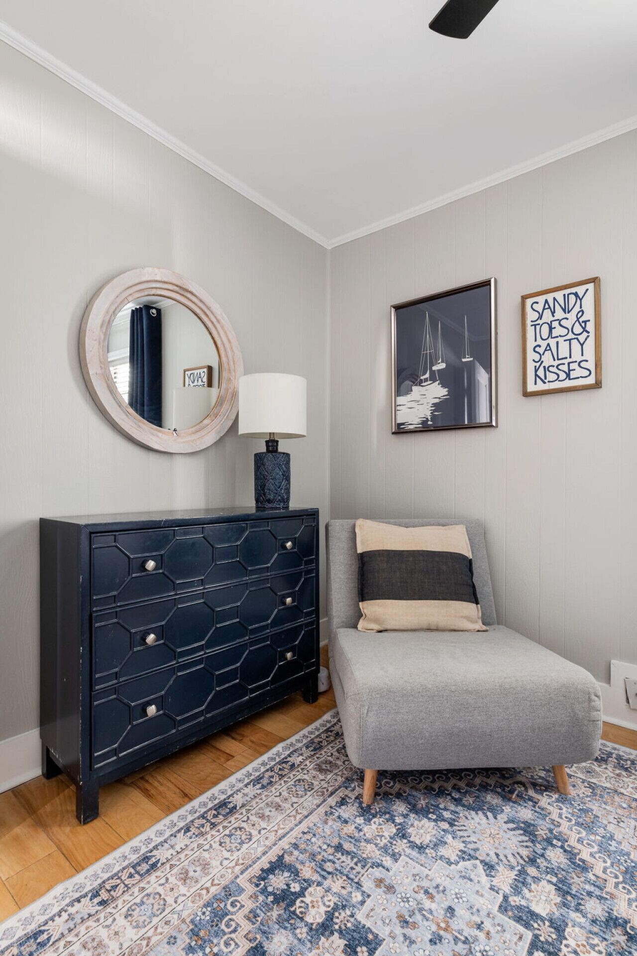A bedroom with a grey sofa, a dark blue dresser, a round mirror, a framed picture, and a patterned rug.