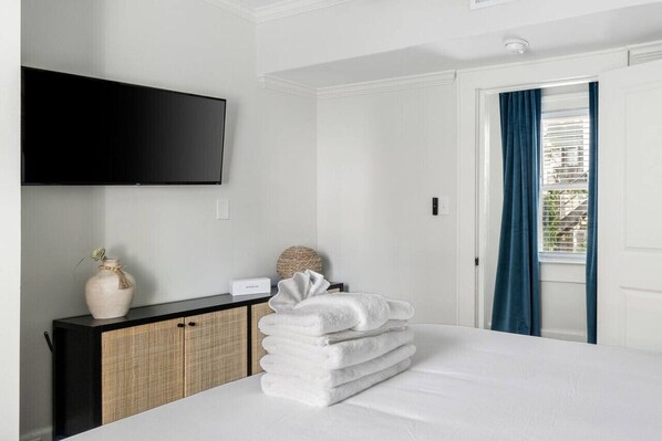 Une chambre d’hôtel avec un lit, un téléviseur à écran plat fixé au mur, une table de chevet avec un vase et une lampe, et une fenêtre avec des rideaux bleus.