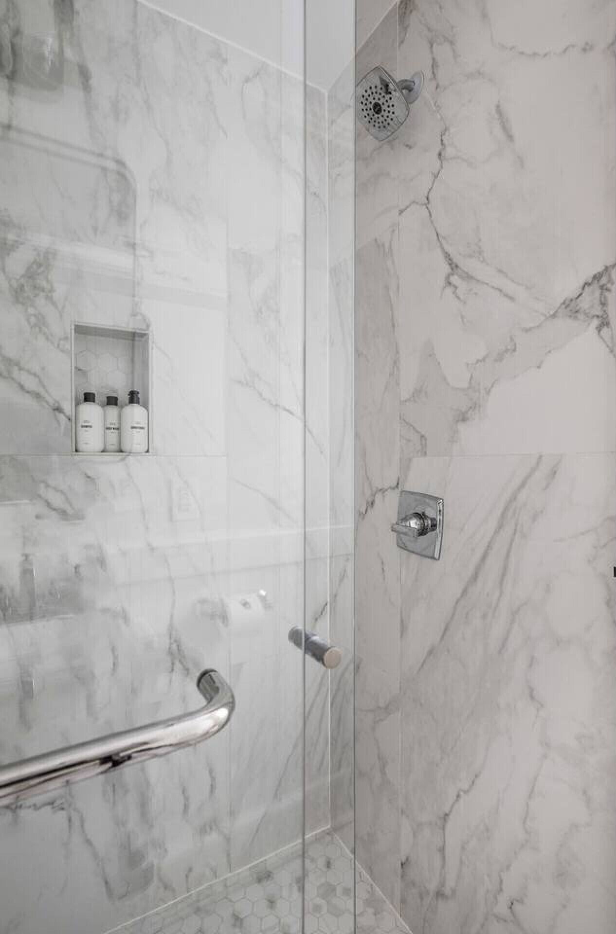 A modern shower with a glass door, marble walls, and a handrail.