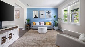 A living room with a blue accent wall, a grey sofa, a white armchair, a round coffee table, and a flat-screen TV on a white cabinet. - The Boroughs on Maxwell - 3 Bedroom (North Charleston)