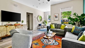 A modern living room with a grey sofa, a coffee table, and a TV. There is a large window with plants and a view of the outside. - Stylish 3BR Near Downtown CHS (North Charleston)