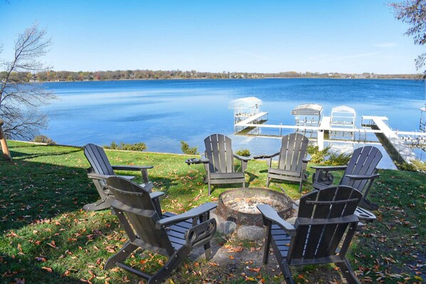 A lakeside seating area with Adirondack chairs and a fire pit, overlooking a dock and calm water.