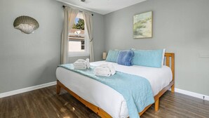 A bedroom with a bed, a ceiling fan, a window with curtains, a wall-mounted lamp, and a painting. - Folly Beach - 2 Blocks to Beach - Sleeps 6! (Folly Beach)