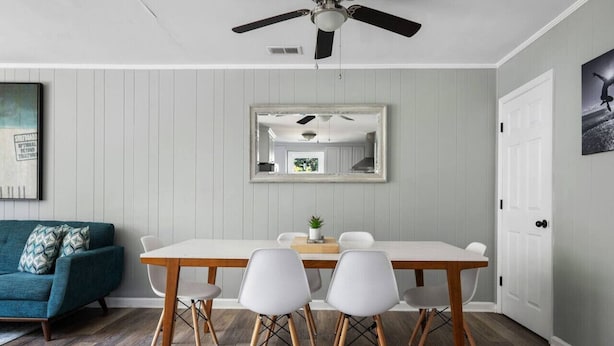 Un comedor con una mesa blanca, cuatro sillas, un ventilador de techo y un espejo grande en la pared.